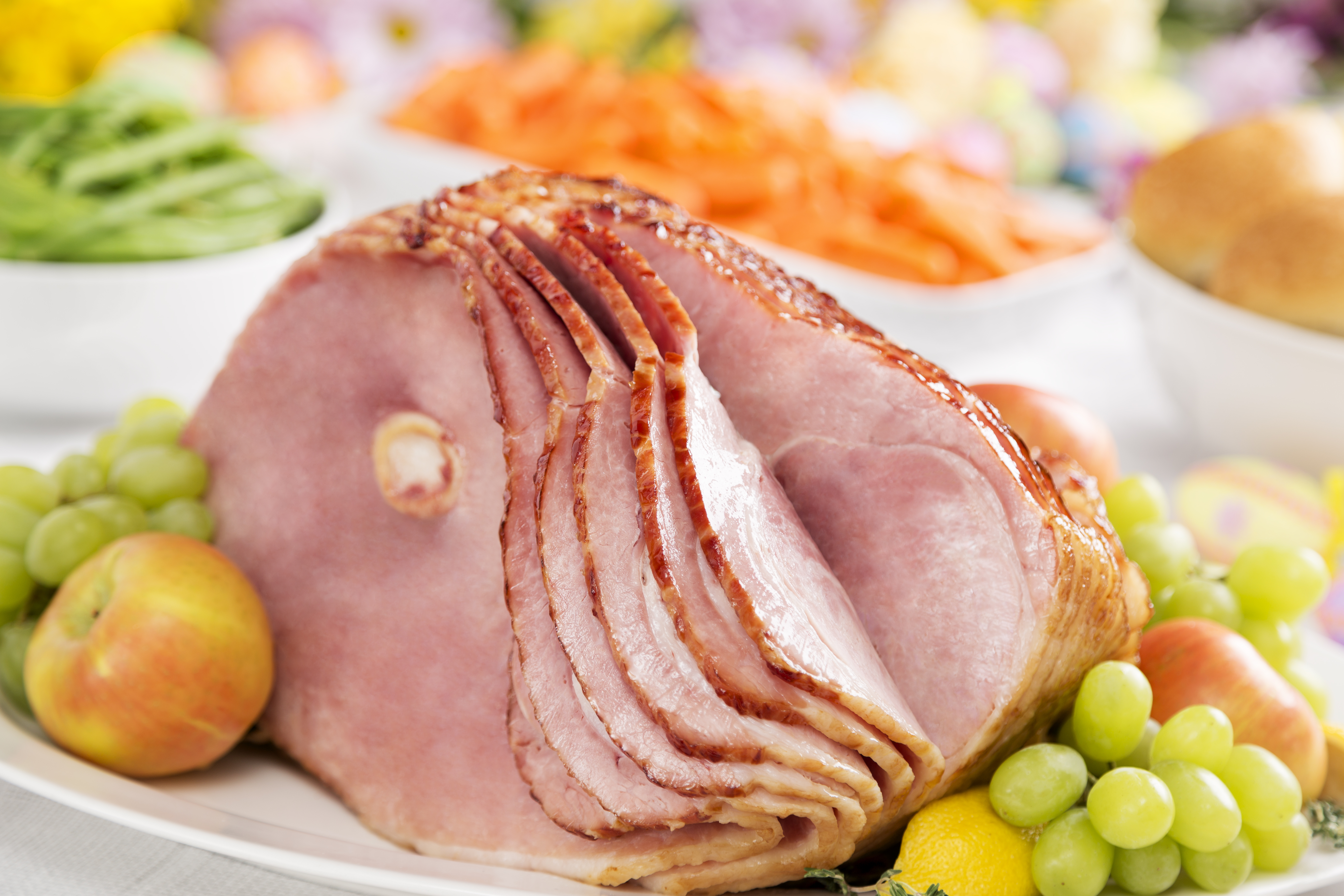 sliced bone-in ham on white plate with apples and grapes on it and blurred veggies in white bowls in background